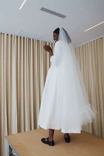 A model wearing a white bridal dress with long sleeves and a square neckline standing on a wooden table
