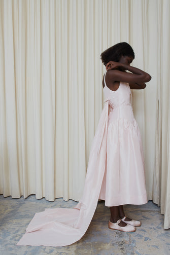 A model wearing a pale pink, long pleated train wedding dress with a V-neck backless design and a waist accentuated with seams. The dress has a drop waisted skirt and a concealed zipper at the side.