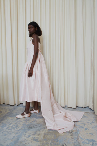 A model wearing a pale pink, long pleated train wedding dress with a V-neck backless design and a waist accentuated with seams. The dress has a drop waisted skirt and a concealed zipper at the side.