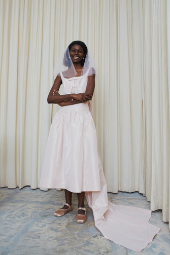 A model wearing a pale pink, long pleated train wedding dress with a V-neck backless design and a waist accentuated with seams. The dress has a drop waisted skirt and a concealed zipper at the side.