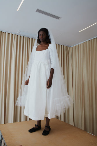 A model wearing a white bridal dress with long sleeves and a square neckline standing on a wooden table