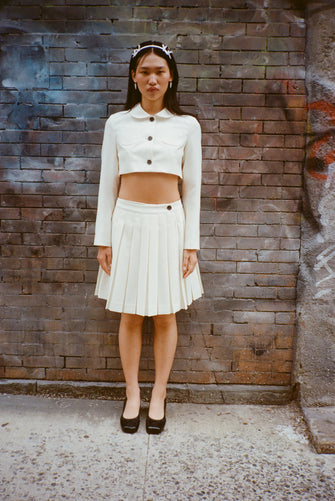 Cream pleated skort with matching cropped jacket on a model against a brick wall 
