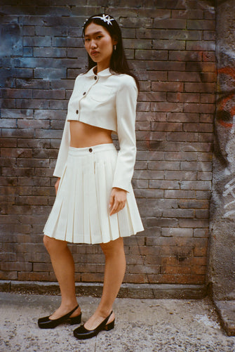 Cream pleated skort with matching cropped jacket on a model against a brick wall 