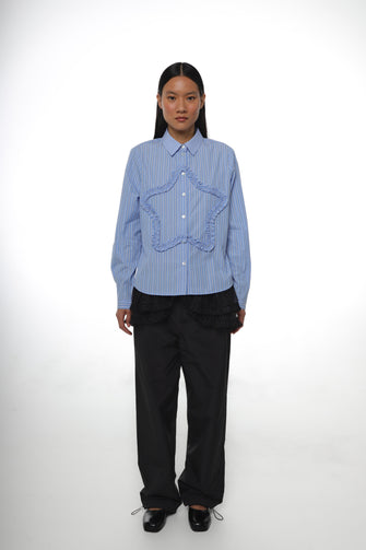 A woman wears a long-sleeved, striped light blue shirt with a ruched star detail at the front and embroidery at the chest pocket with black track pants and black shoes against a white background