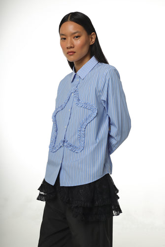 A woman wears a long-sleeved, striped light blue shirt with a ruched star detail at the front and embroidery at the chest pocket with black track pants against a white background