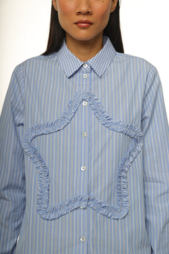 A woman wears a long-sleeved, striped light blue shirt with a ruched star detail at the front and embroidery at the chest pocket against a white background