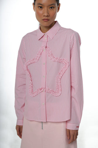 A woman wears a long-sleeved pink striped shirt with a ruched star detail at the front and embroidery at the chest pocket with a pink skirt against a white background