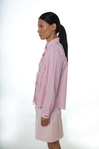 A woman wears a long-sleeved pink striped shirt with a ruched star detail at the front and embroidery at the chest pocket with a pink skirt against a white background