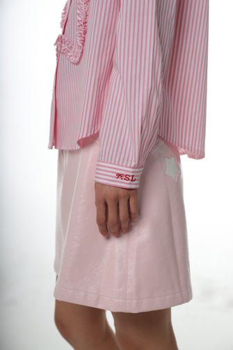 A woman wears a long-sleeved pink striped shirt with a ruched star detail at the front and embroidery at the chest pocket with a pink skirt against a white background