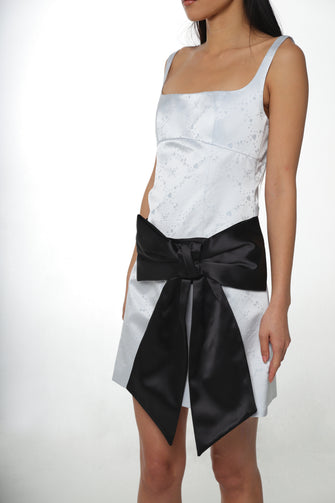 A woman wears a blue satin dress with a black adjustable sash bag with a bow design, displayed against a plain background.
