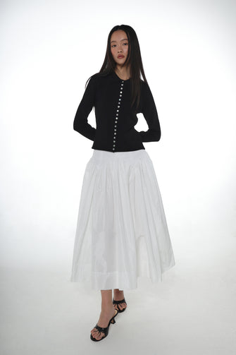 A woman wears  long sleeved black cardigan with mother of pearl heart, star and flower buttons with a white skirt and black heels